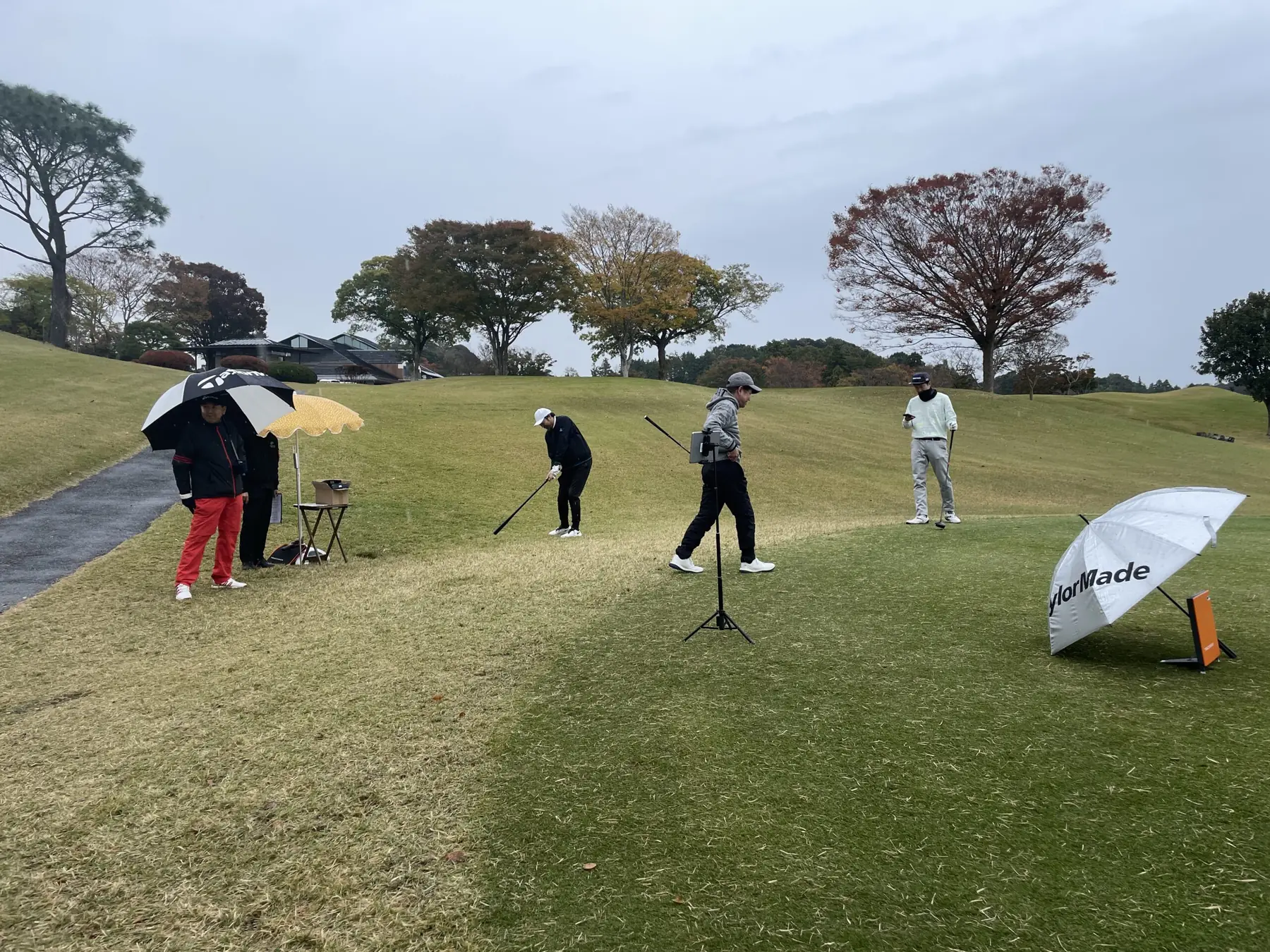 紅葉の残るゴルフコースのティーで打つゴルファーと、計測機器や傘を囲む参加者。