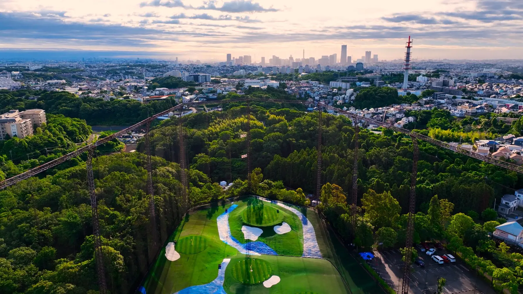 HANZ GOLF CLUBのレンジを上空から見た様子。森に囲まれたフェアウェイとネットの支柱、奥に横浜の高層ビル群。