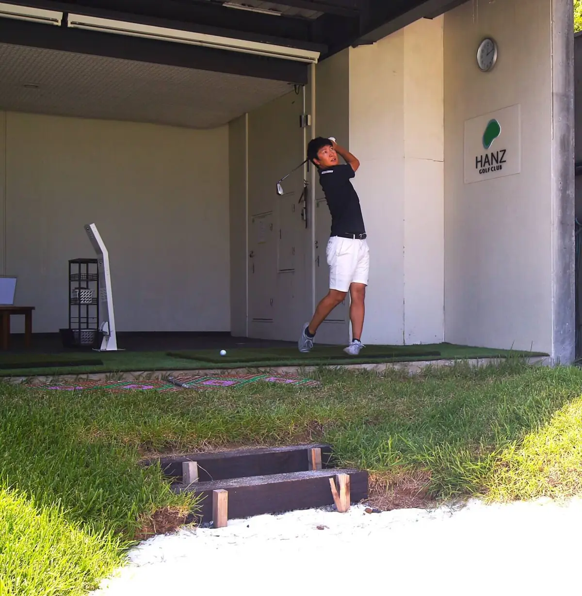 HANZ GOLF CLUBのロゴが掲げられた屋根付き打席でアイアンを振り抜く男性。