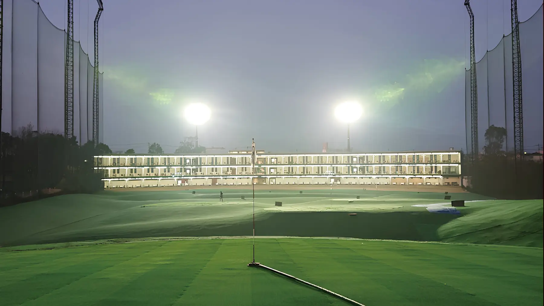 夜のHANZ GOLF CLUBのレンジ。外野から見た照明付きの3階建て打席棟と2基の投光器、手前にピンの立つ芝。