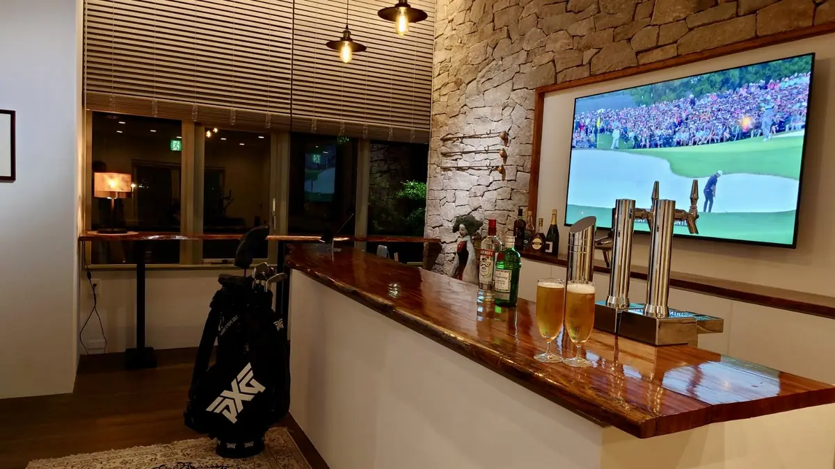 一枚板のバーカウンターに置かれたビール2杯とビールサーバー。石壁の大型テレビにゴルフ中継、手前にキャディバッグ。